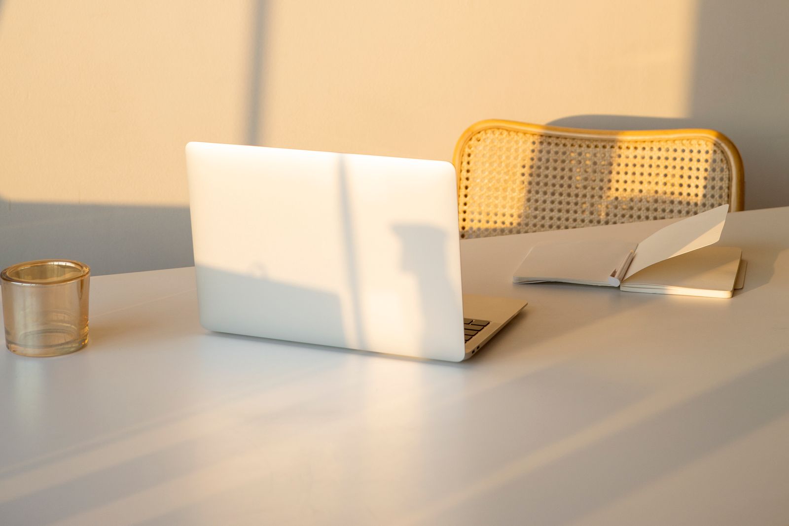 A sunlit workspace with a laptop, an open notebook and a cane chair